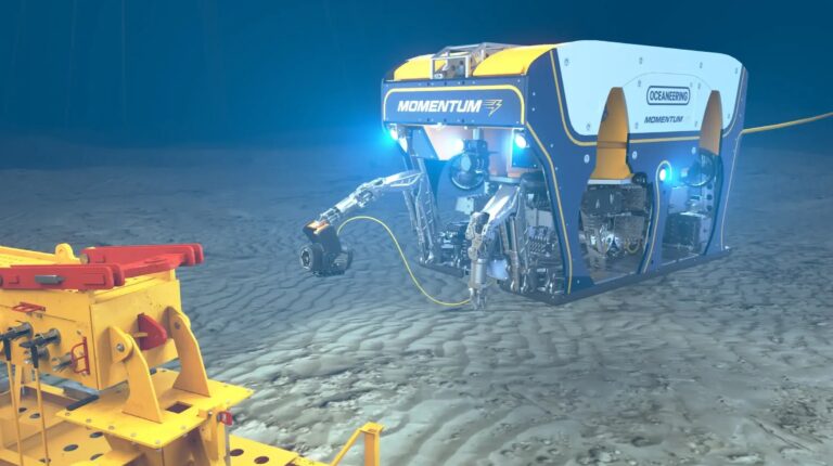 The Oceaneering Momentum electric work-class ROV carrying out a job underwater. it is square in shape; has blue, yellow and grey colouring and branding; several lights, arms and a camera at the front. The seabed is visible below and another underwater vehicle is in front of it.